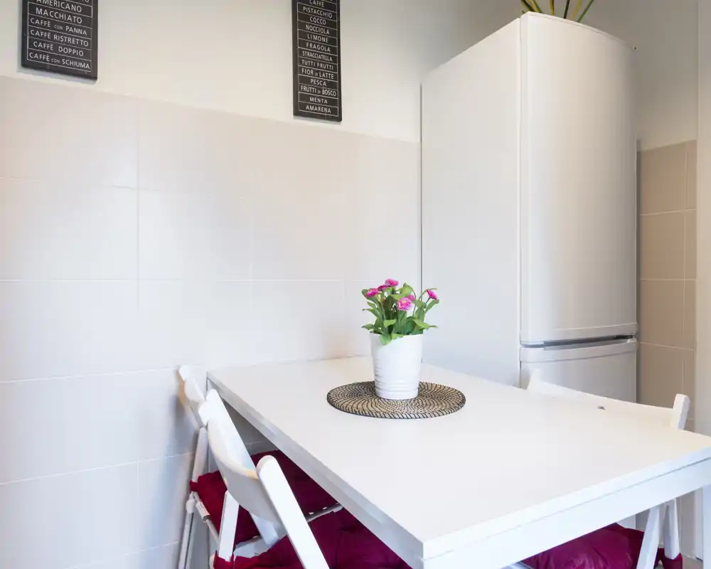 Accogliente angolo pranzo luminoso con tavolo bianco, sedie pieghevoli con cuscini rossi e una pianta al centro — perfetto per pasti informali in un angolo cucina compatto.