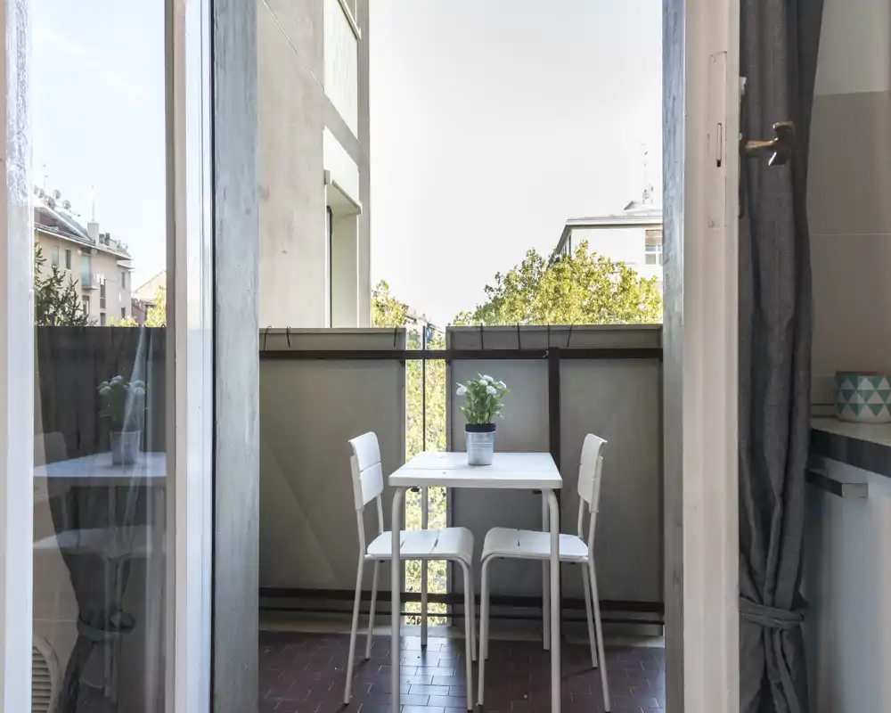 Piccolo balcone con un tavolo bianco compatto e due sedie, vaso di piante al centro e vista sui palazzi vicini — perfetto per il caffè mattutino.