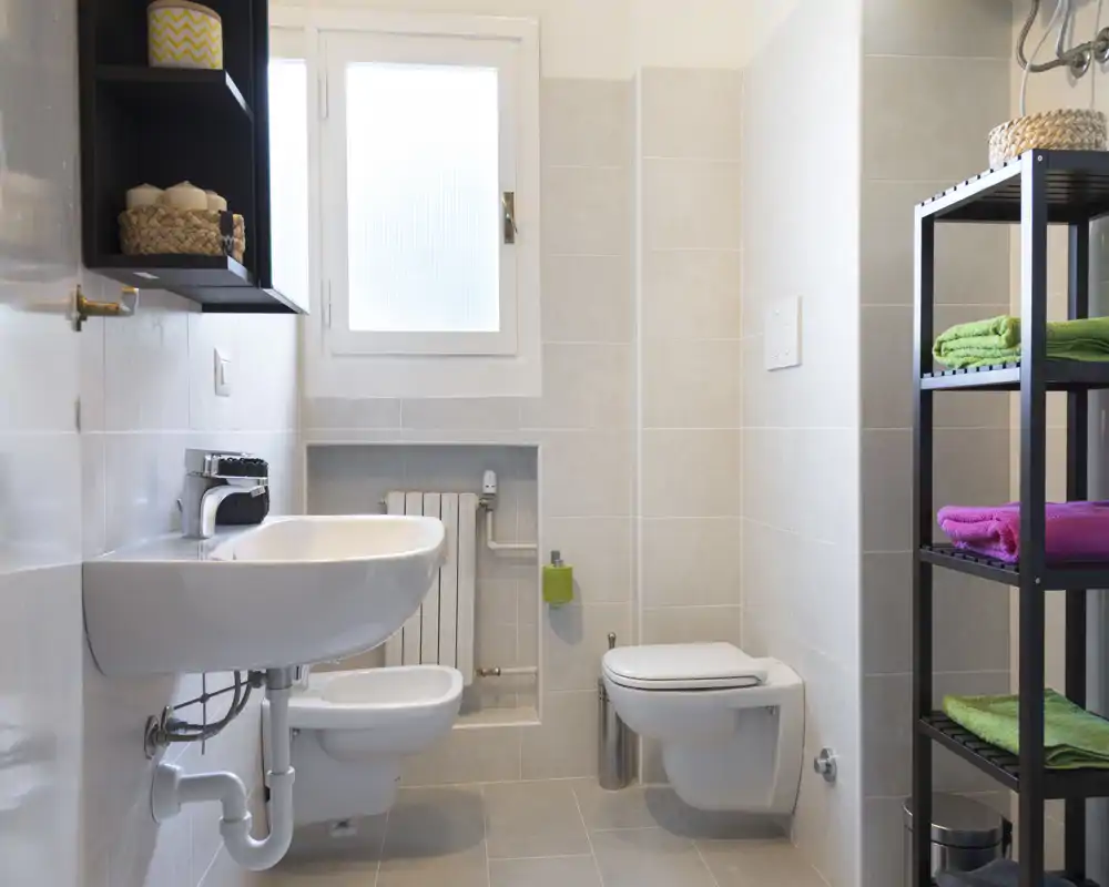 Bagno moderno e luminoso con wc sospeso e bidet, lavabo con tubature a vista e scaffale con asciugamani colorati.