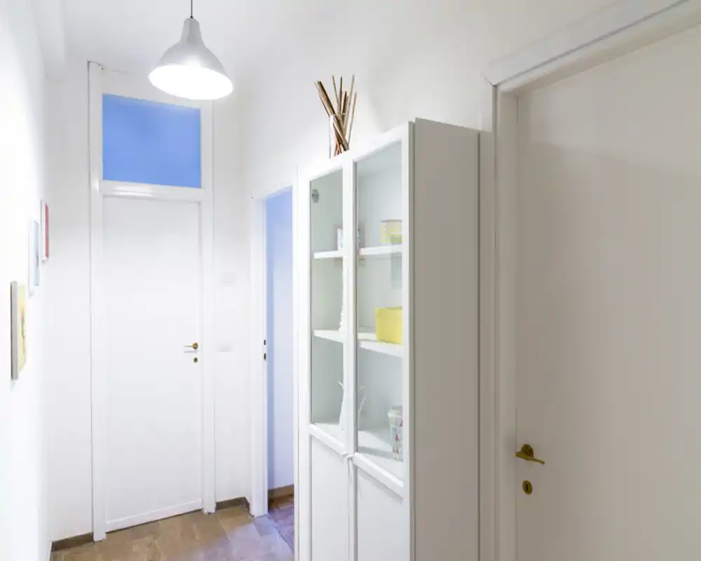 Ingresso luminoso e stretto con pareti bianche, lampada a sospensione e porta chiusa; una vetrina bianca offre spazio contenitivo e scaffalatura decorativa.