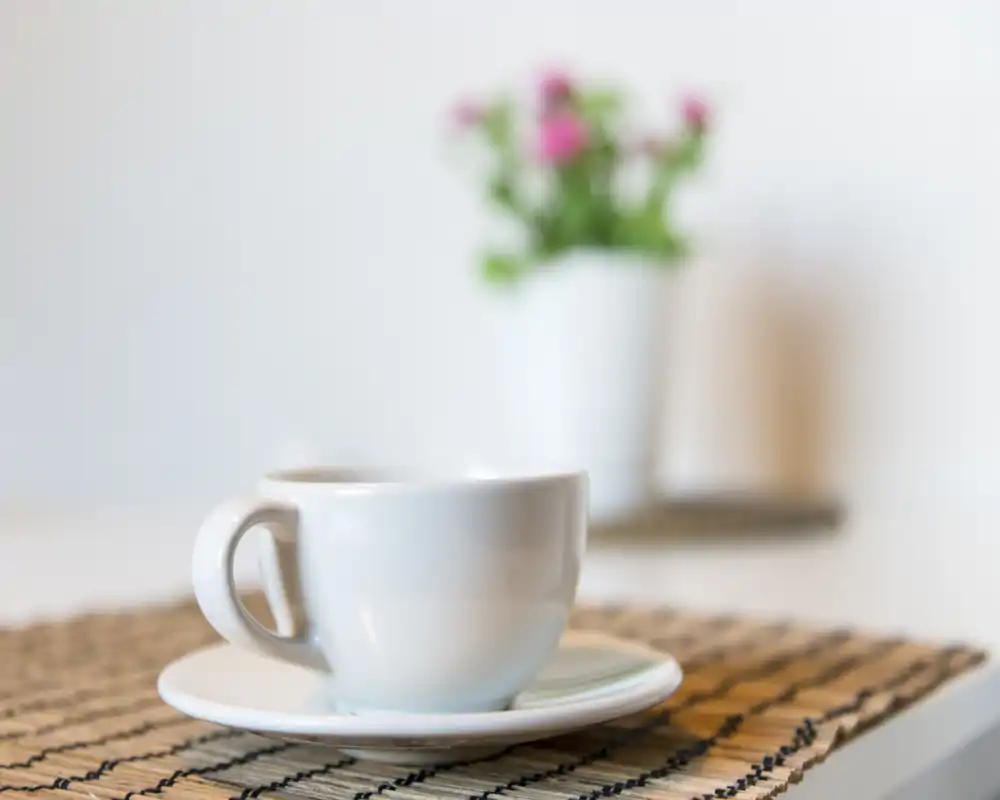 Primo piano di una tazza da caffè bianca con piattino su un tovaglietta, sfondo morbido e sfocato; ambiente luminoso e minimal, perfetto per annunci stile colazione o caffetteria.