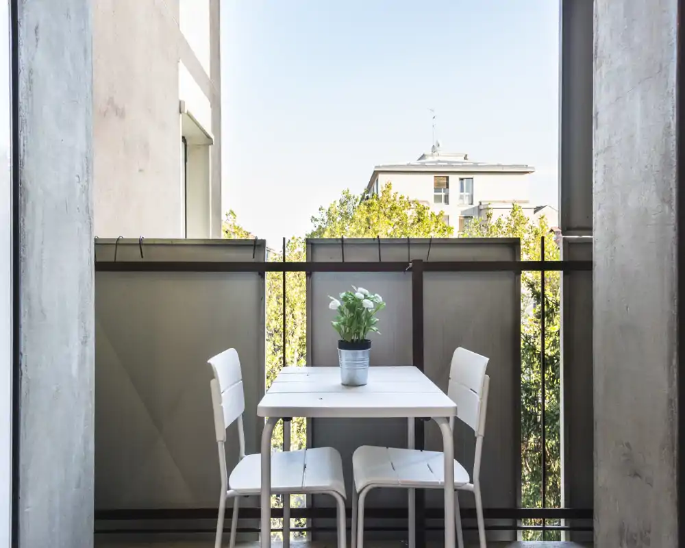 Balconcino soleggiato con un piccolo tavolo bianco e due sedie, ideale per un caffè mattutino; una pianta in vaso aggiunge un tocco di verde e i pannelli garantiscono privacy.