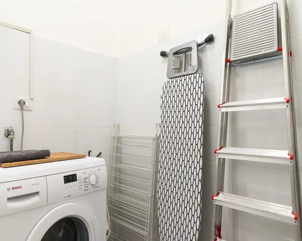 Angolo lavanderia/utility compatto con lavatrice a carica frontale, asse da stiro, stendibiancheria e scaletta in alluminio; ambiente pulito, ben illuminato e funzionale.