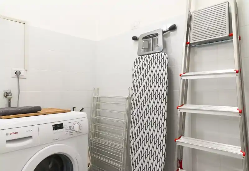 Rincón de lavandería/utility compacto con lavadora de carga frontal, tabla de planchar, tendedero y escalera de aluminio; limpio, bien iluminado y funcional.