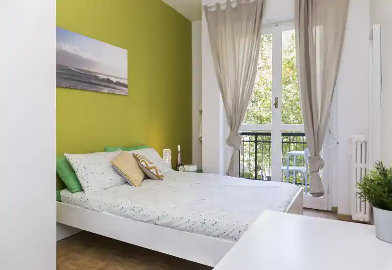 Bright, modern bedroom with a double bed, decorative cushions and a green accent wall; large window/door opens to a small balcony letting in abundant natural light.