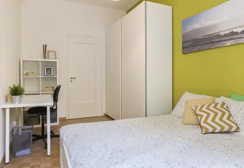 Bright, modern bedroom with a large bed dressed in patterned linens, decorative cushions and a green accent wall; includes wardrobe and a small desk for work.