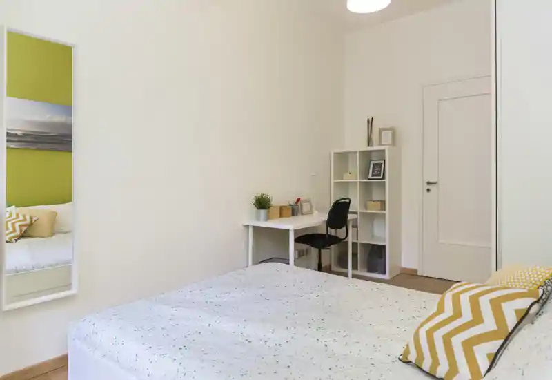 Bright, minimalist bedroom with a neatly made double bed, patterned cushions and a small workspace with shelving — ideal for a comfortable short or long stay.
