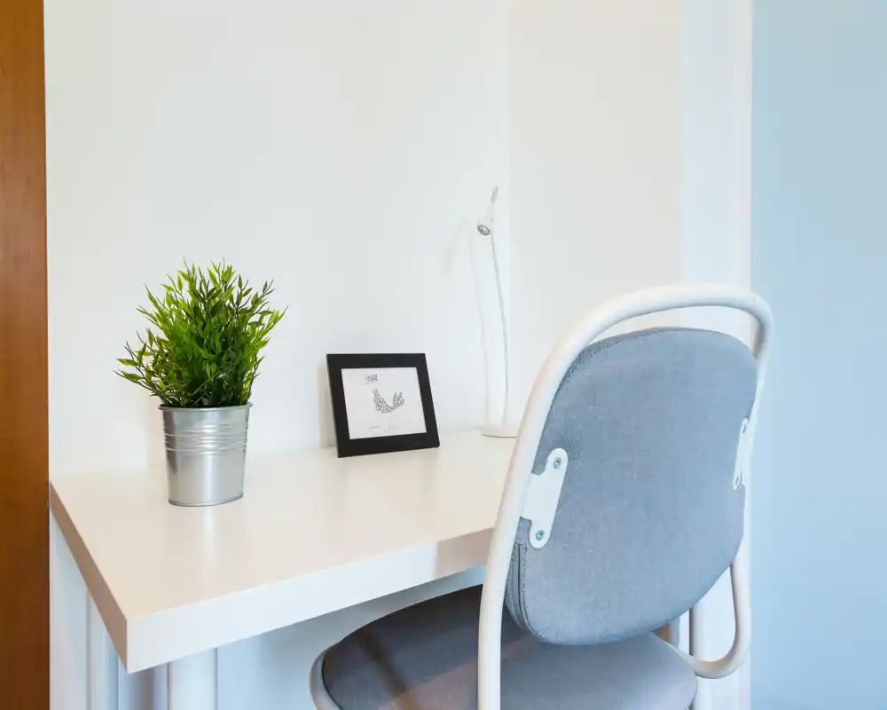 Rincón de trabajo luminoso y minimalista con escritorio blanco, silla gris acolchada, pequeña planta y marco decorativo — ideal para concentrarse en un espacio doméstico reducido.