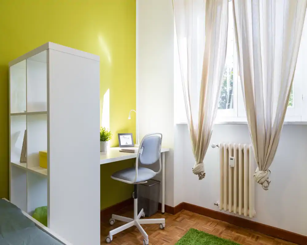 Rincón de dormitorio con escritorio compacto, silla y estantería; la luz entra por cortinas translúcidas y se ve parte de la cama en el borde izquierdo.