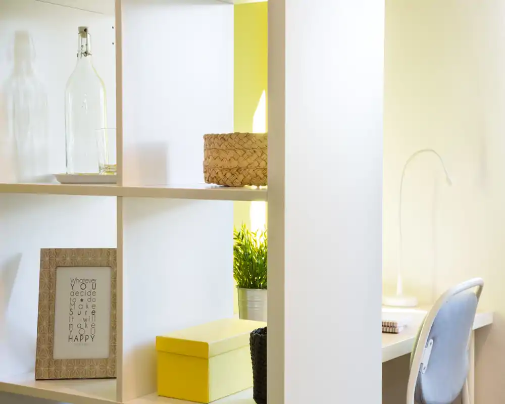 Rincón de estar luminoso y minimalista con estantería decorativa, planta en maceta, cesta trenzada y un pequeño escritorio — ideal como espacio funcional en un piso compacto.