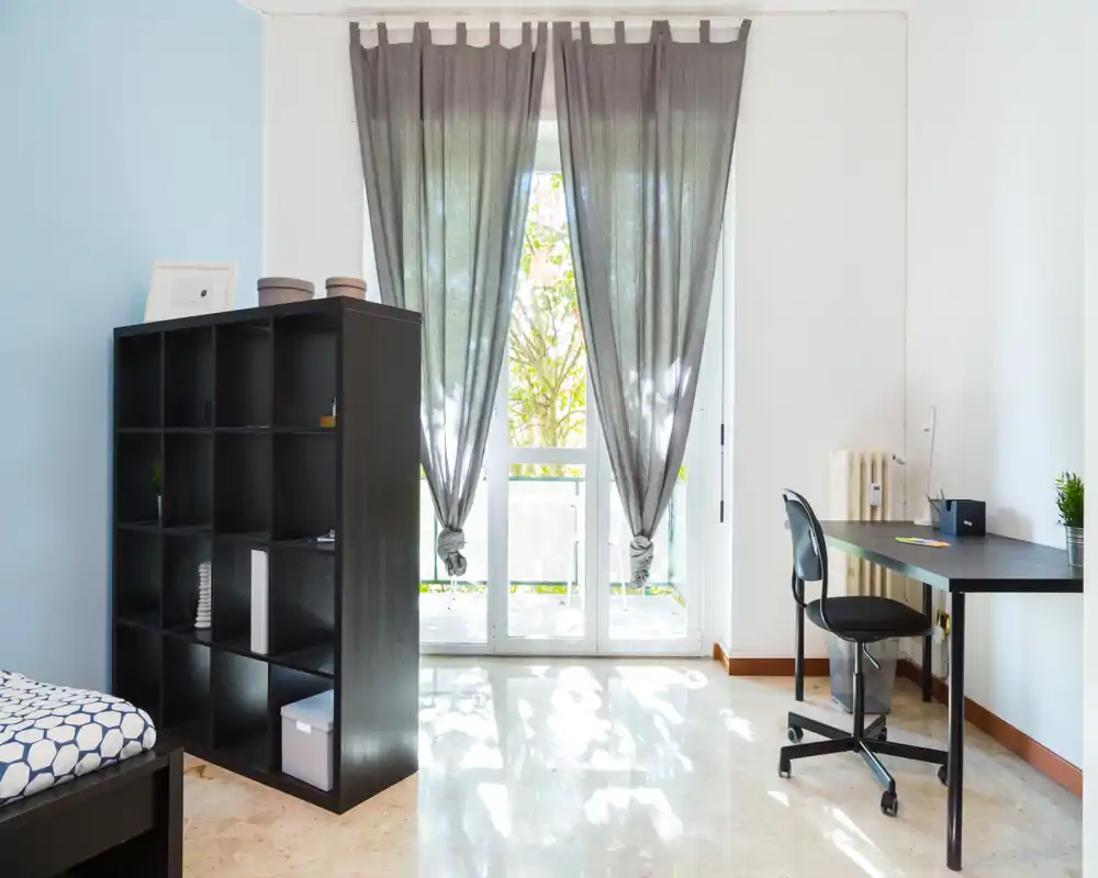 Dormitorio luminoso y ordenado con cama individual parcialmente visible, estantería negra como separador y escritorio junto a la puerta del balcón con cortinas semitransparentes.