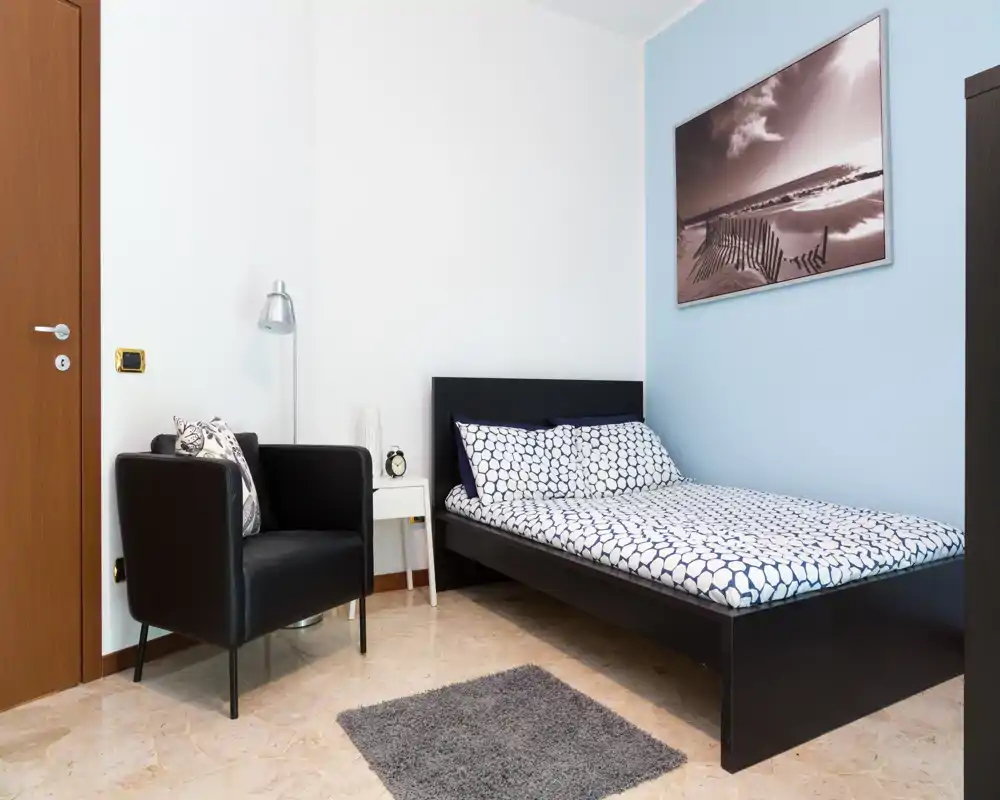 Dormitorio moderno y luminoso con cama doble, ropa de cama estampada, un sillón negro y mesita de noche sencilla — espacio minimalista y bien decorado, ideal para alquileres vacacionales.