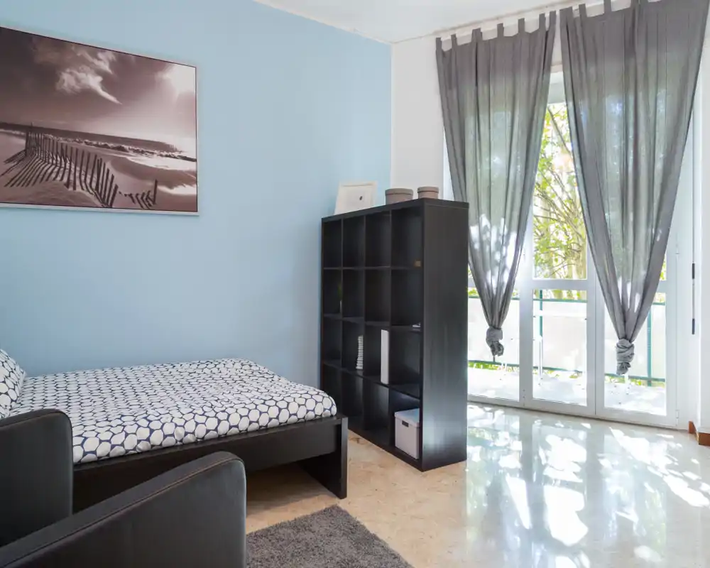 Dormitorio luminoso y ordenado con una cama individual estampada, estantería modular y grandes ventanas que dejan entrar mucha luz natural — paredes azules y decoración moderna y minimalista.