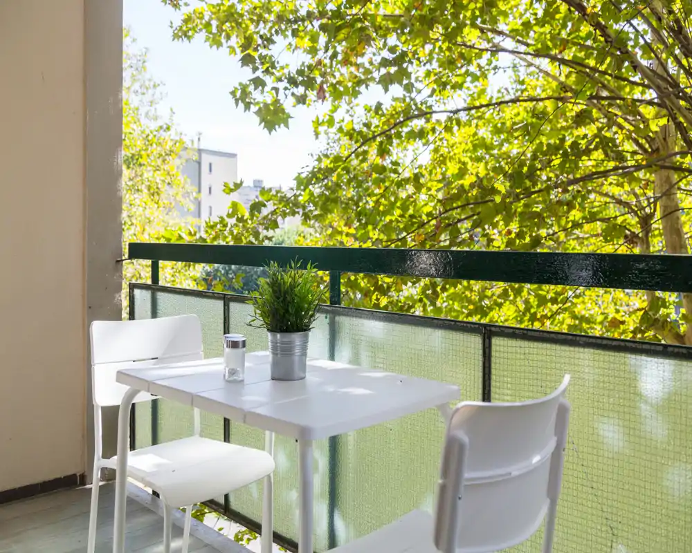 Balcón soleado con una mesa blanca, dos sillas y una planta en maceta — un rincón luminoso y con vegetación para tomar café o leer.