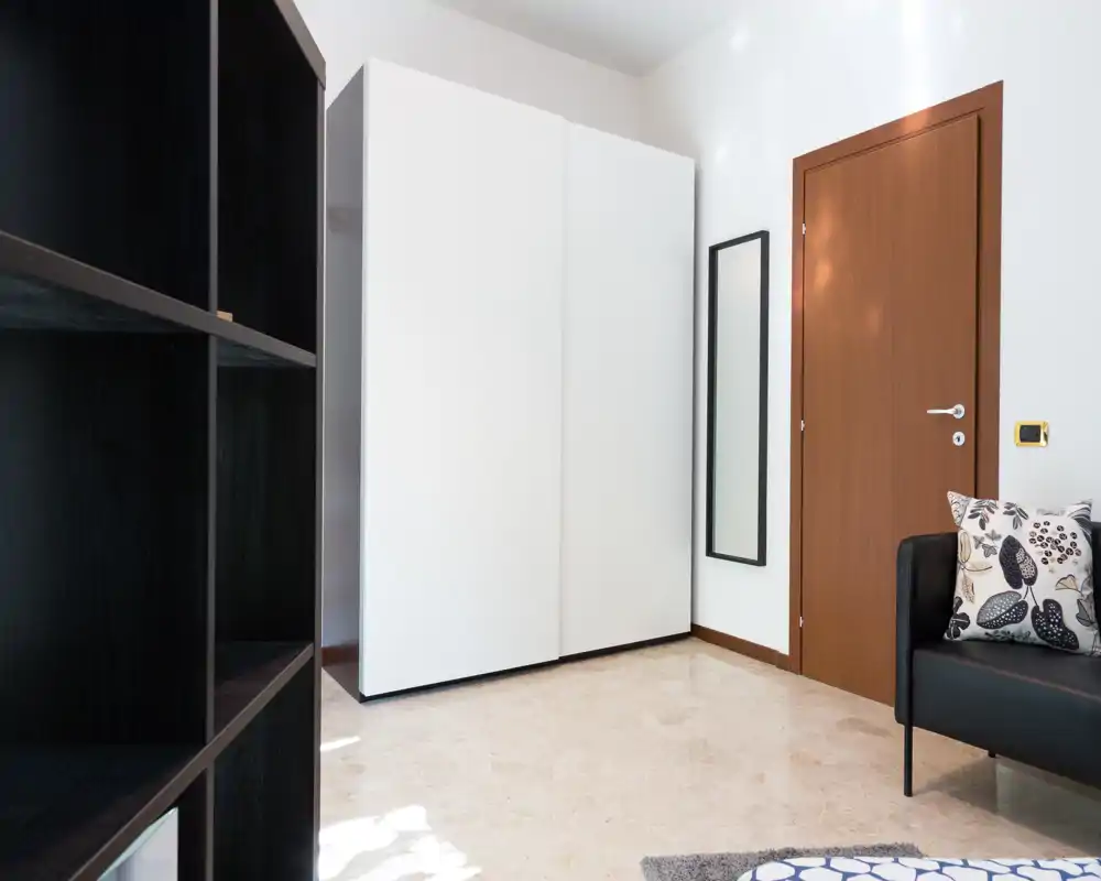 Zona de estar minimal y luminosa con sofá compacto, cojines decorativos, estantería y un gran armario blanco: estilo moderno y tonos neutros.