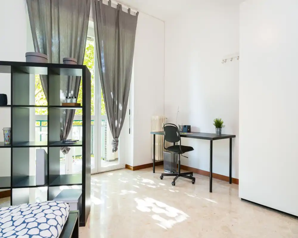 Dormitorio luminoso y minimalista con cama individual, estantería abierta y un pequeño escritorio junto a la puerta del balcón que aporta mucha luz natural.