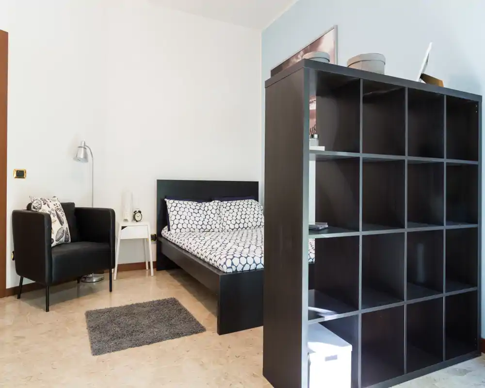Dormitorio bien presentado con cama doble, ropa de cama con estampado, un sillón acogedor y una estantería separadora que crea una división sutil. Luminoso y ordenado, perfecto para anuncios de alquiler que destaquen la distribución y el estilo.