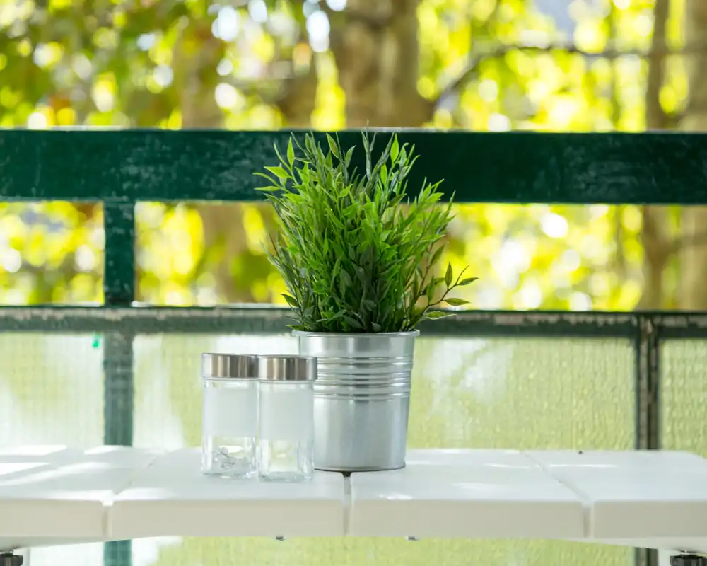 Escena luminosa de balcón con una pequeña maceta metálica con planta y dos recipientes de vidrio sobre una mesa blanca; el fondo de hojas sugiere un exterior.