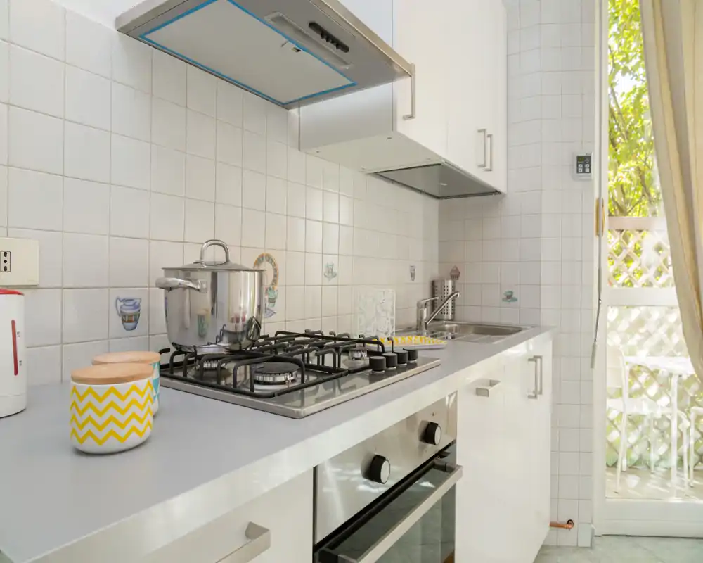 Cocina moderna y luminosa con placa de gas, horno empotrado, fregadero y tarros decorativos; la luz natural de la puerta aporta una sensación fresca y acogedora.
