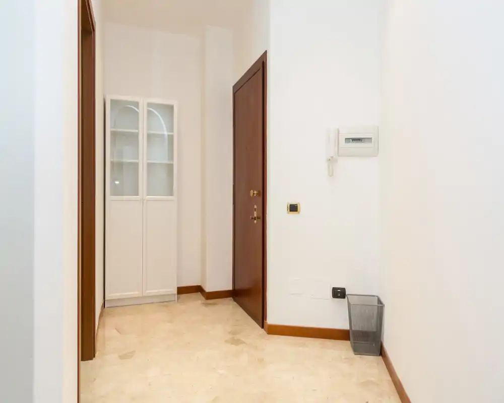 Entrada luminosa y ordenada con puerta de madera, paredes blancas y un armario estrecho — espacio de llegada limpio y acogedor.
