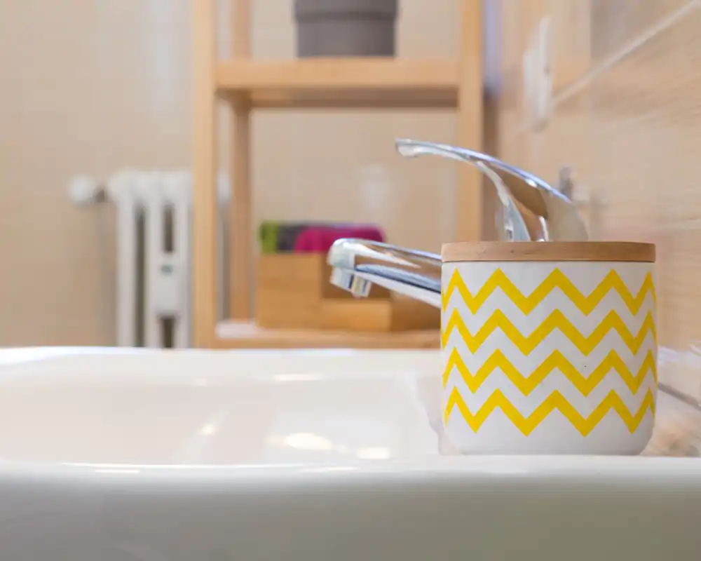 Primer plano de un lavabo de baño moderno con grifo cromado y un portacepillos decorativo con zigzag amarillo en el borde del lavabo, con azulejos y accesorios limpios.