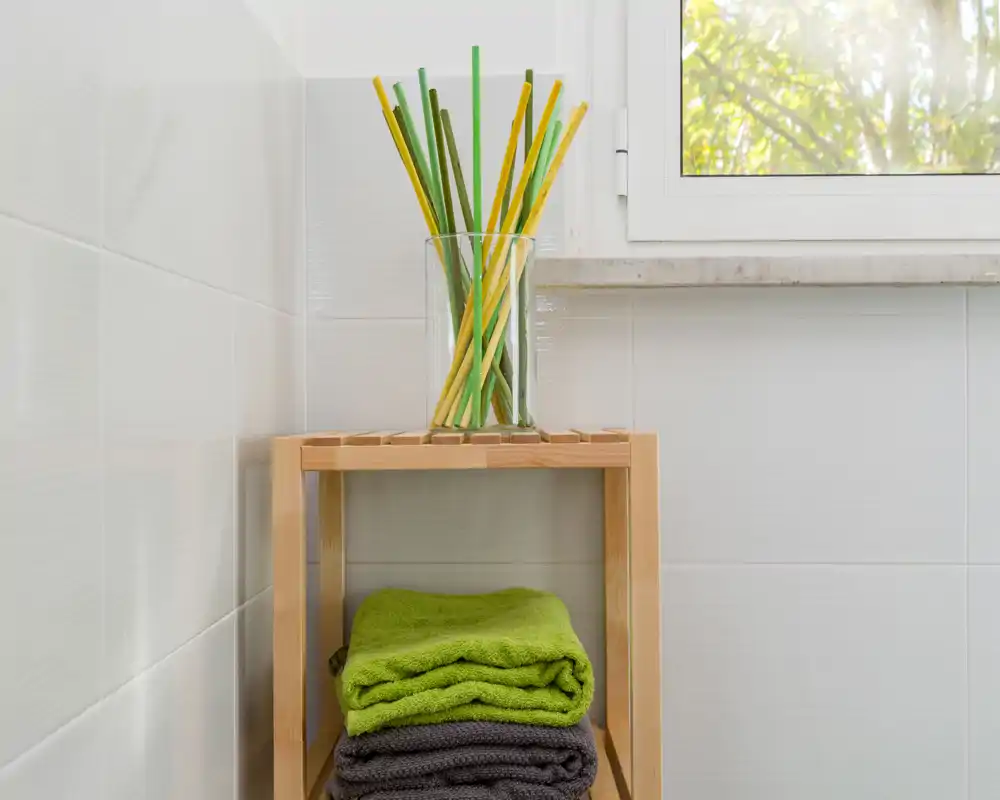 Detalle del baño: estantería de madera con toallas dobladas y un jarrón de vidrio sobre azulejos blancos bajo una ventana luminosa.