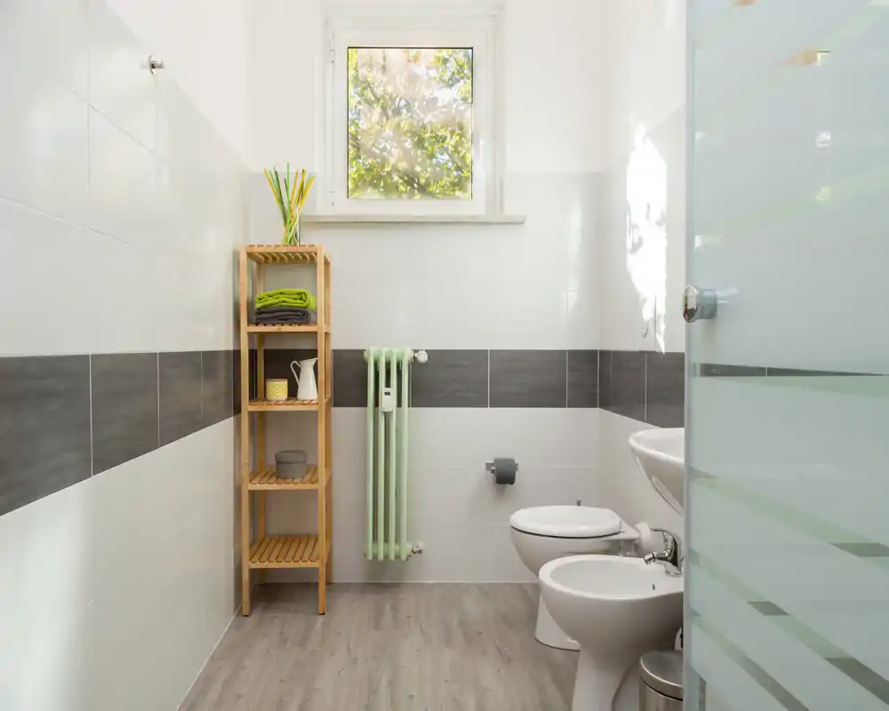 Baño moderno y luminoso con bidé y WC, estantería de madera clara y ventana que proporciona luz natural.