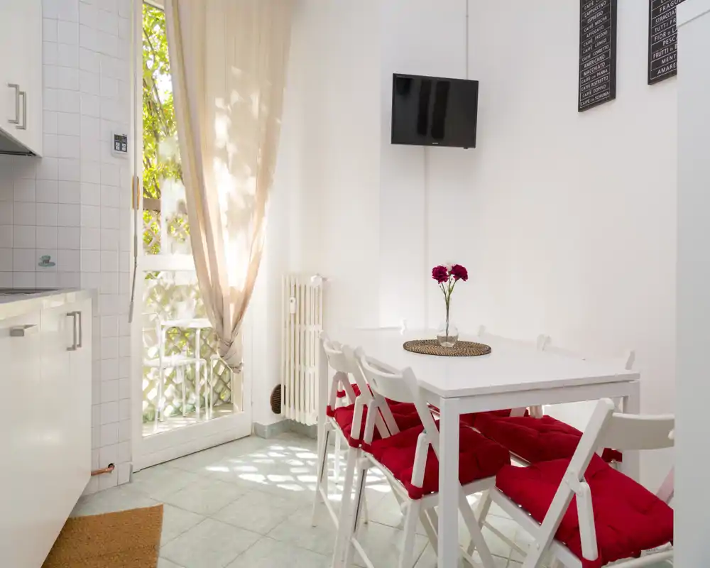 Área de comedor compacta y luminosa con mesa y sillas blancas con cojines rojos, luz natural por la puerta de cristal y un pequeño jarrón en el centro — ideal para comidas acogedoras.
