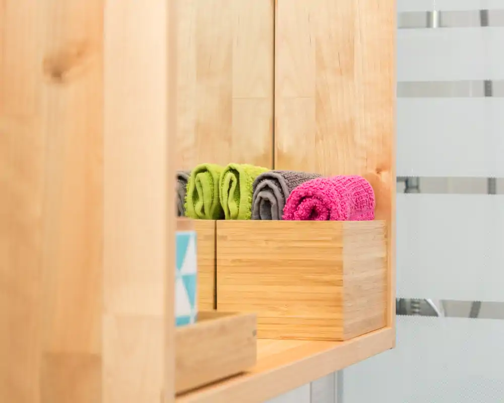 Primer plano de una estantería de baño con toallas de colores enrolladas en una caja de madera, junto a un vidrio esmerilado. Detalle limpio y minimalista.