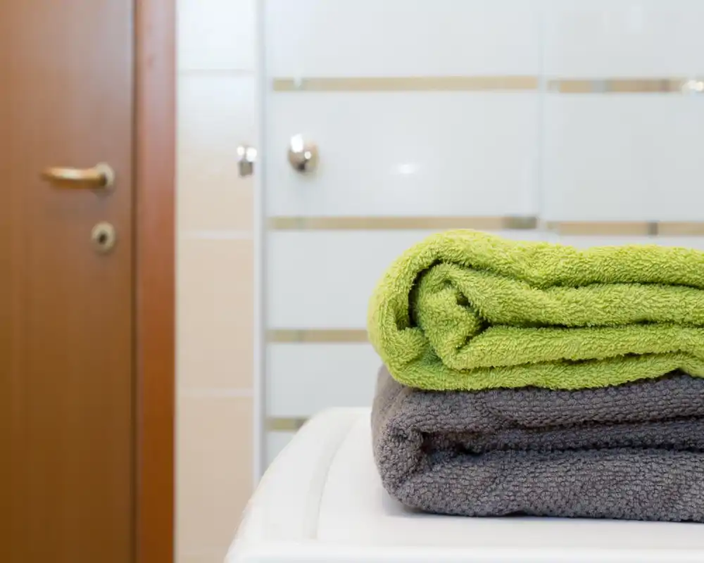 Toma nítida en primer plano de toallas dobladas sobre una lavadora o superficie del baño, con puerta de ducha satinada y puerta de madera al fondo — detalle de baño ordenado y bien iluminado.