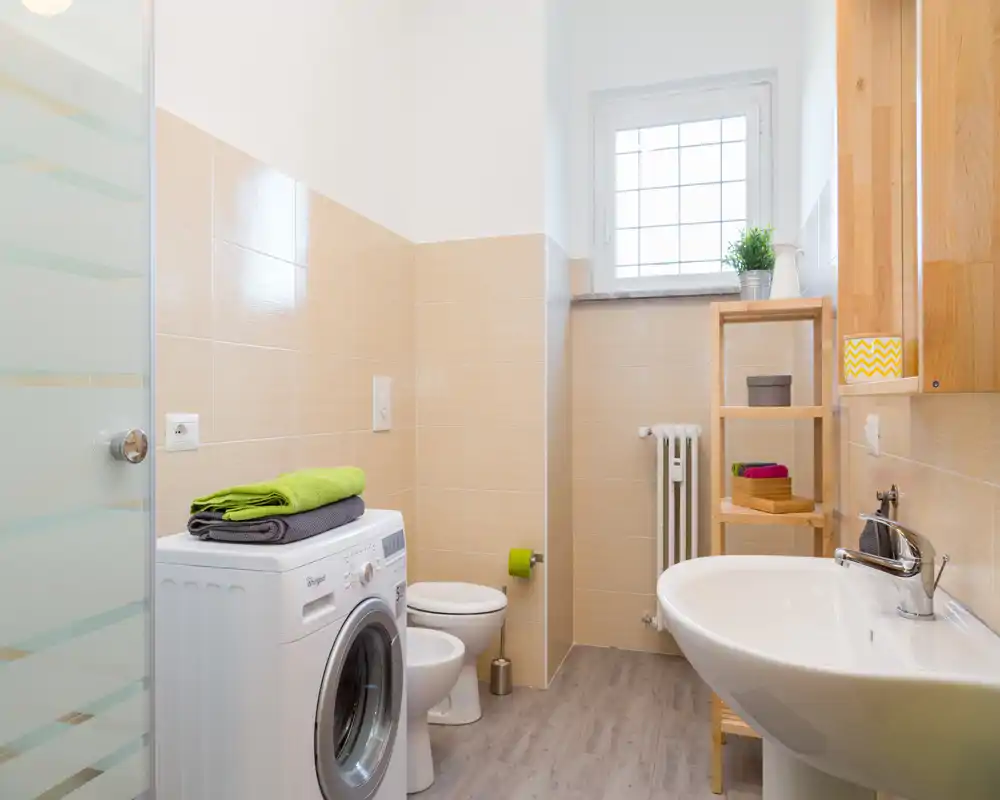 Baño moderno y luminoso con lavabo, inodoro y lavadora; azulejos beige y una pequeña estantería ofrecen almacenamiento práctico.