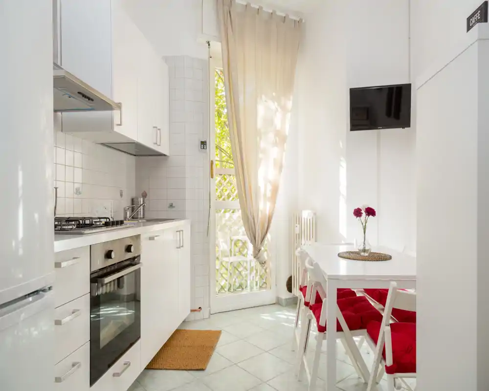 Cocina compacta y luminosa con muebles blancos, cocina de gas y un pequeño comedor con sillas con cojines rojos; luz natural que entra por una cortina ligera.