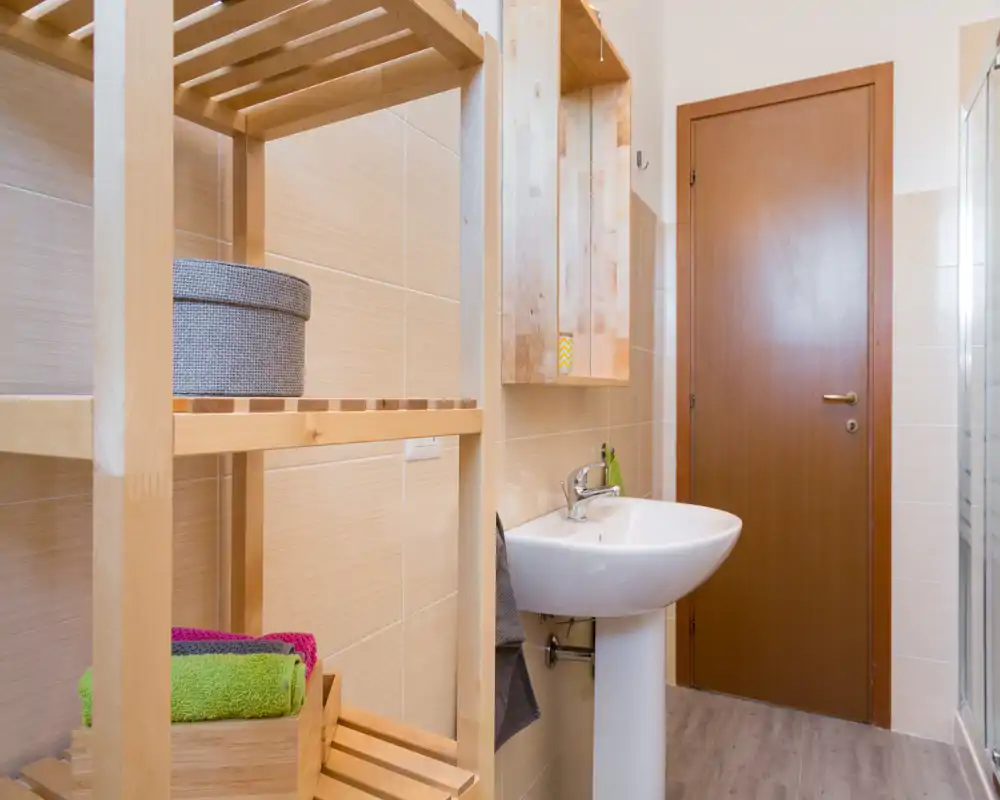 Baño luminoso y ordenado con lavabo de pedestal, estanterías de madera y un elemento de ducha de vidrio visible al borde de la imagen.