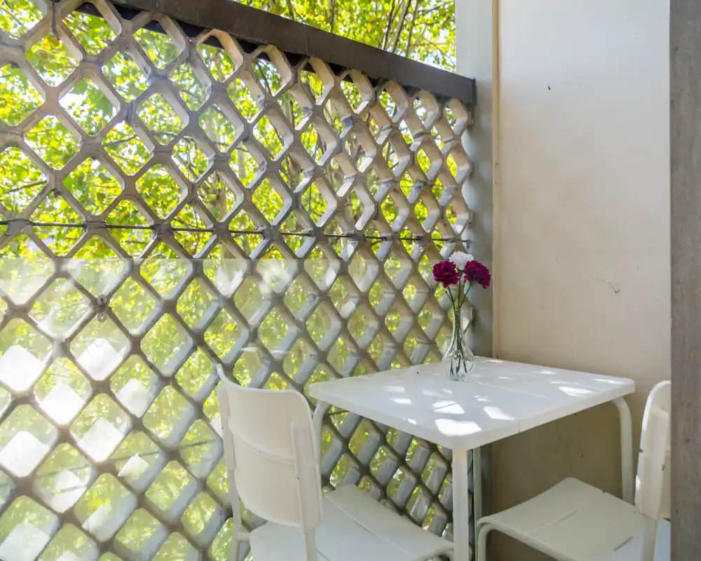 Pequeño balcón soleado con mesa y sillas blancas, celosía decorativa y jarrón con flores — ideal para el café de la mañana.