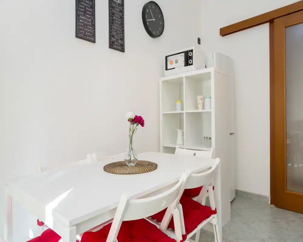 Zona de comedor luminosa y sencilla con mesa blanca, cuatro sillas con cojines rojos y una estantería pequeña; flores decorativas y un reloj de pared aportan una sensación acogedora y ordenada.