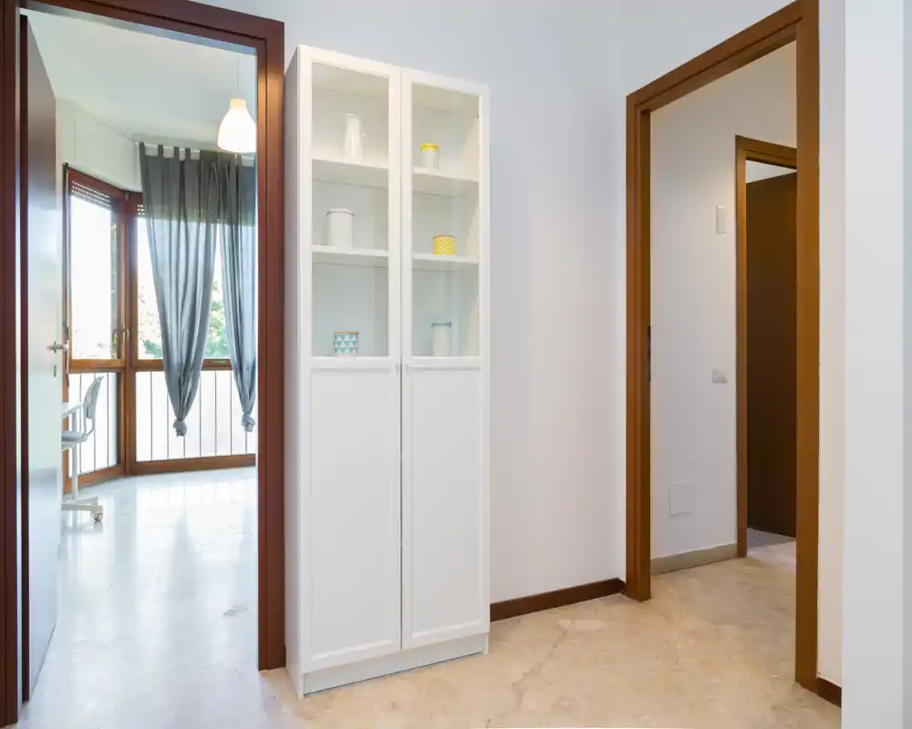 Entrada/pasillo luminoso y minimalista con un armario blanco y puertas que conducen a habitaciones contiguas; suelo pulido y luz natural desde la sala contigua crean una sensación de amplitud.