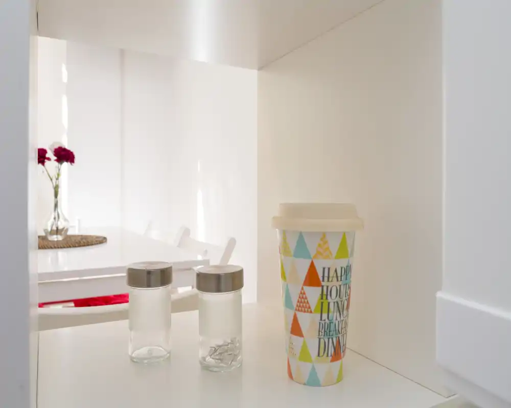 Vista luminosa y minimalista del comedor con mesa y sillas blancas y un jarrón decorativo; ideal para anuncios que resaltan un rincón de comedor moderno y limpio.