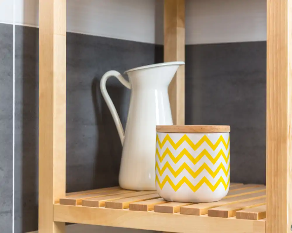 Plano detalle de una estantería decorativa con una jarra de cerámica y un tarro estampado sobre lamas de madera frente a un fondo de azulejos — ideal para mostrar un rincón de balcón o almacenamiento exterior.