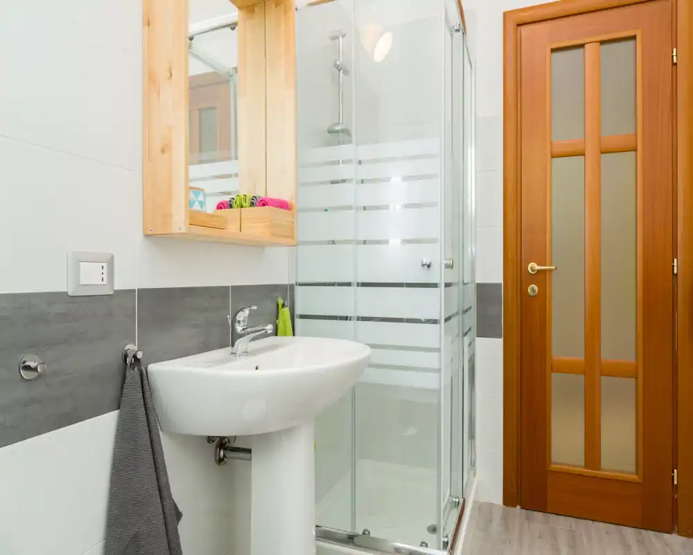 Baño moderno y luminoso con lavabo de columna, mampara de ducha de cristal y puerta de madera; estilo limpio y sencillo, ideal para anuncios.