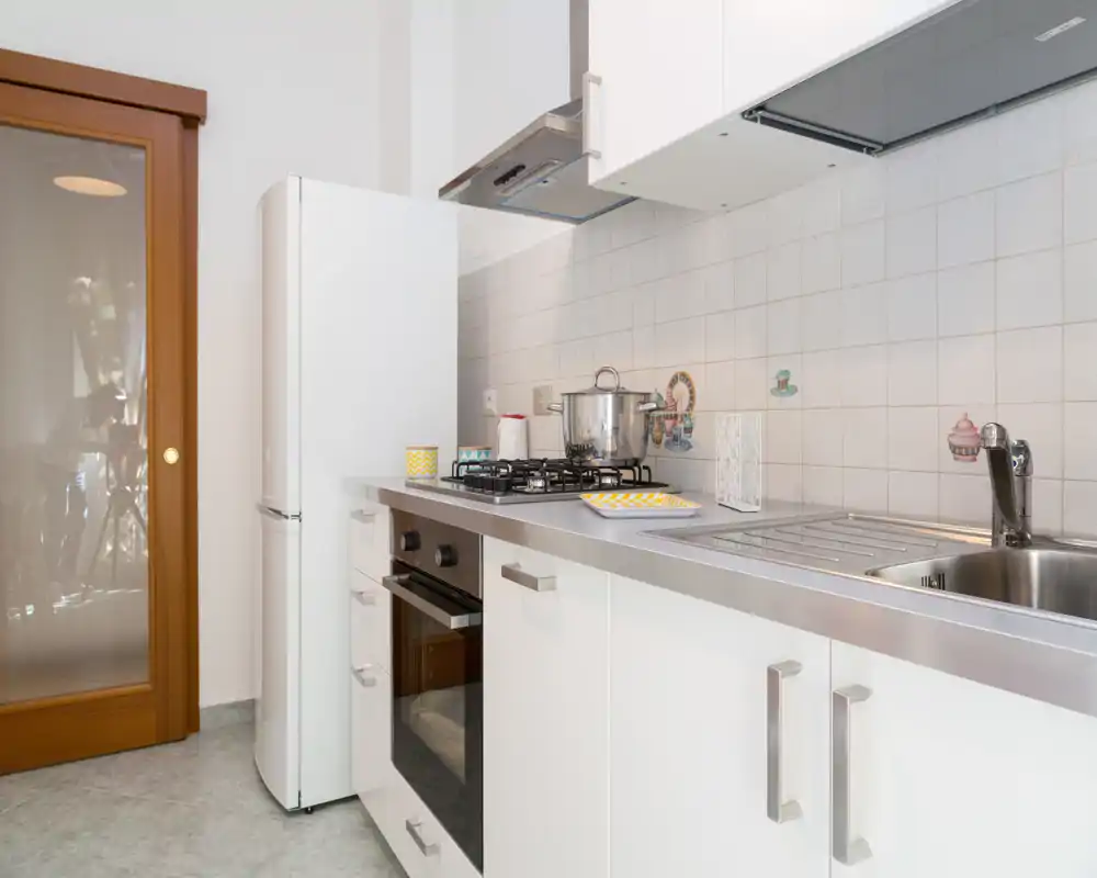Cocina moderna y compacta con armarios blancos, fregadero de acero, placa de gas y horno integrado; limpia, luminosa y ordenada, ideal para alquileres temporales.