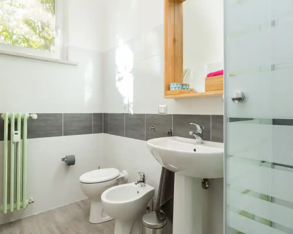 Baño luminoso y limpio con lavabo, bidé, inodoro y mampara de ducha de vidrio; azulejos neutros y espejo con marco de madera para un aspecto moderno y fresco.