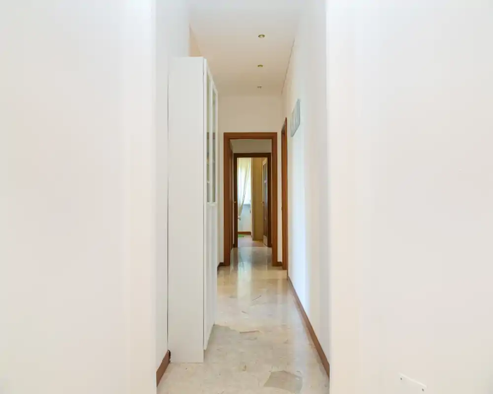 Pasillo luminoso y minimalista con suelo de mármol y focos empotrados que conduce a las habitaciones; paredes blancas limpias que dan sensación de amplitud.