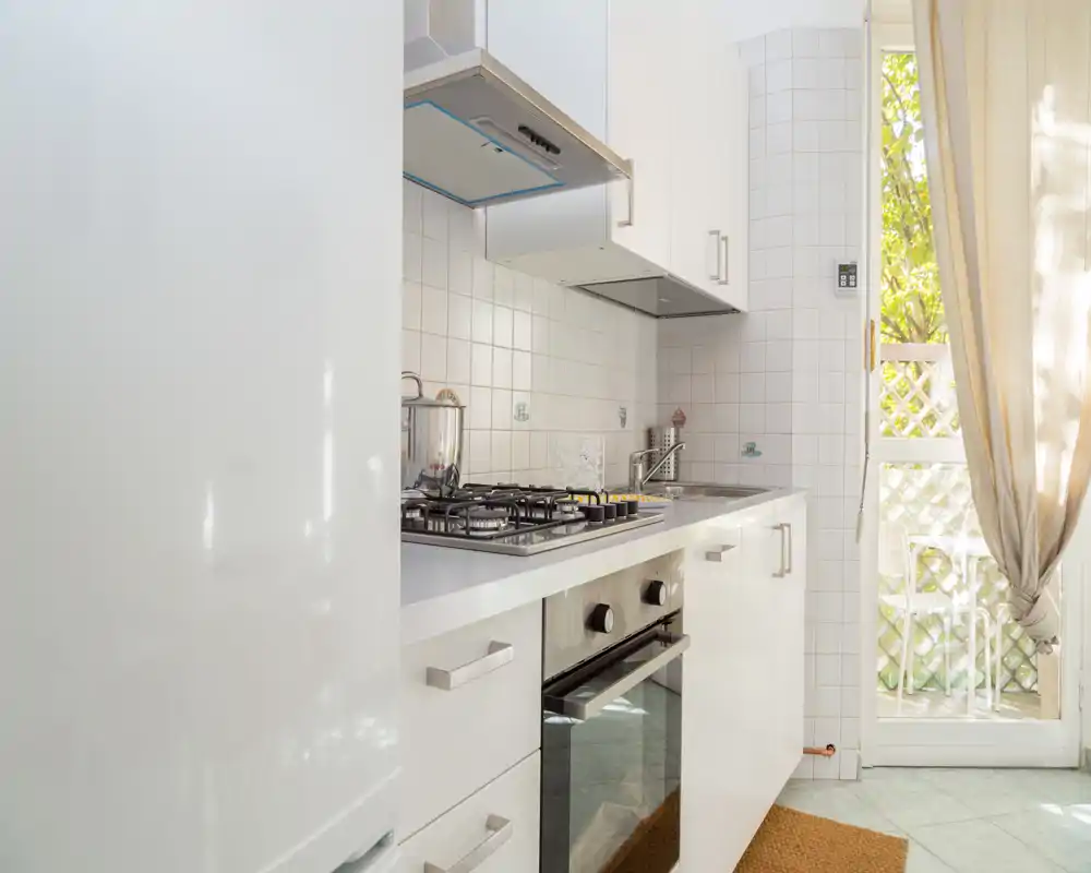 Cocina moderna y luminosa con armarios blancos, placa de gas y horno empotrado; la luz natural desde la puerta y los textiles suavizan el ambiente.