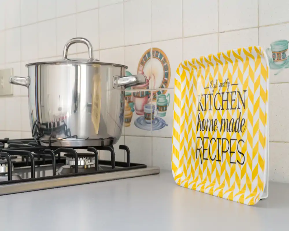 Plano detalle de una encimera de cocina y una cocina de gas con una olla de acero inoxidable y una bandeja decorativa que dice 'Kitchen home made recipes'. Salpicadero alicatado limpio y una vista luminosa y bien presentada del área de cocción.