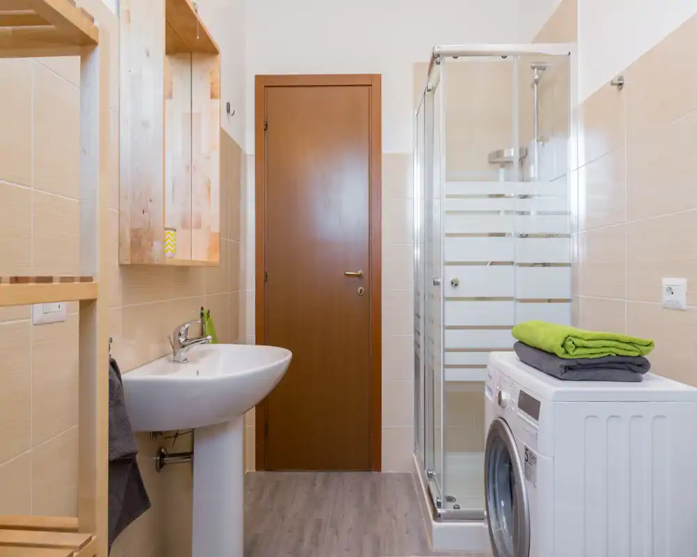 Baño moderno y luminoso con ducha de esquina de cristal, lavabo de pedestal y lavadora; azulejos neutros y detalles de madera crean un espacio limpio y funcional.