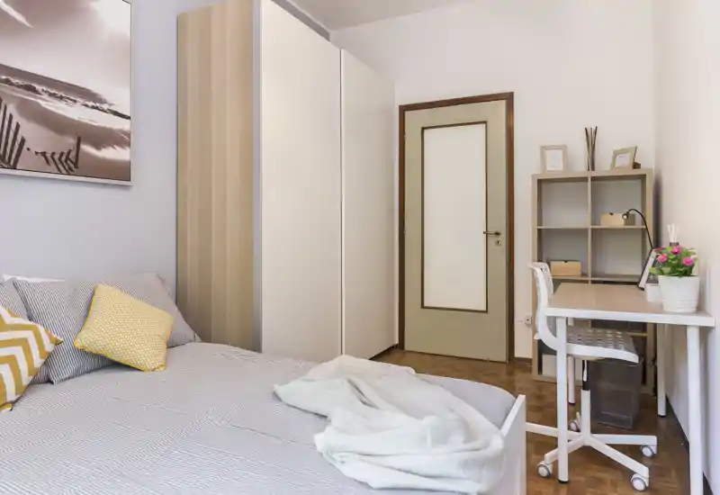 Bright, neatly styled bedroom with a double bed, wardrobe and a compact desk — ideal for a student or professional. Soft textiles and accent cushions create a welcoming atmosphere.