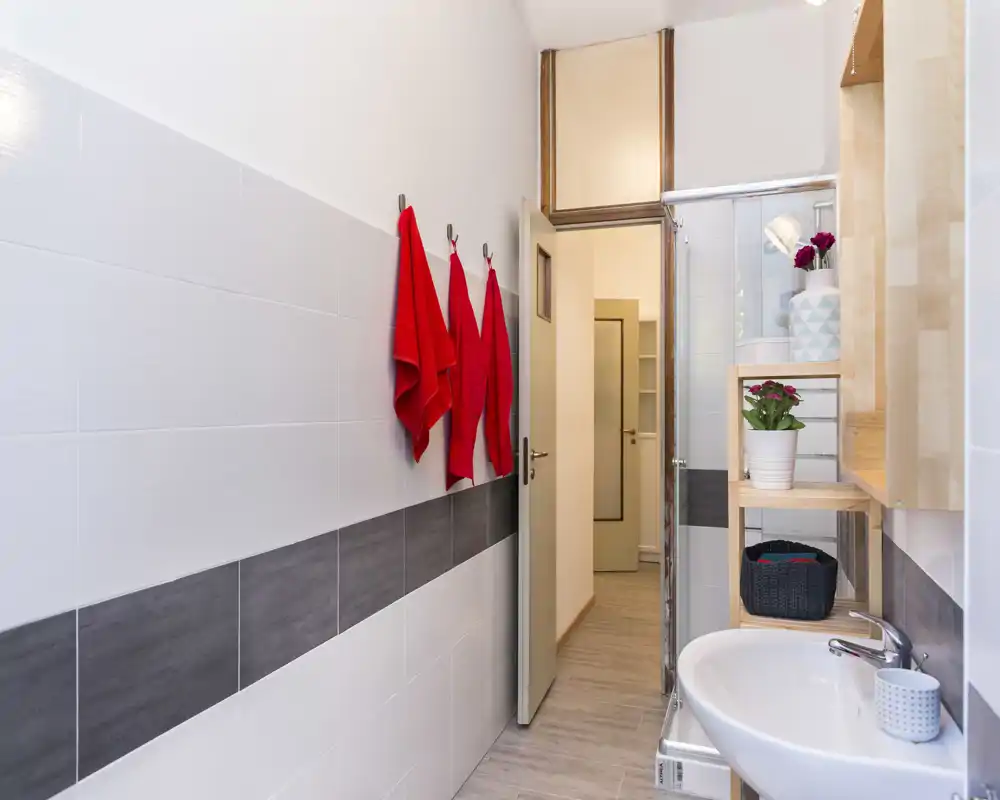 Bright, modern bathroom with white and gray tiled walls, a wall-mounted sink, shelving with decorative plants and towels hung on hooks — simple and clean.