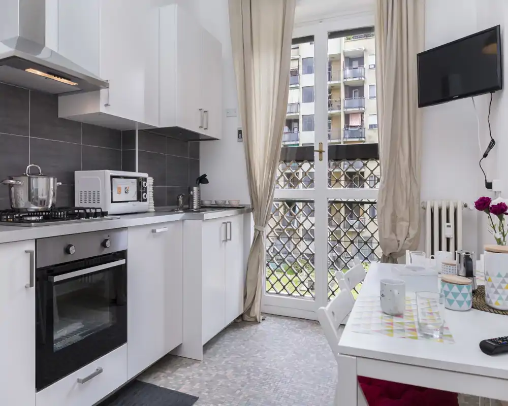 Bright, modern kitchenette with white cabinets, gas hob and oven, small dining table and natural light from a balcony door — practical and well-styled for city stays.