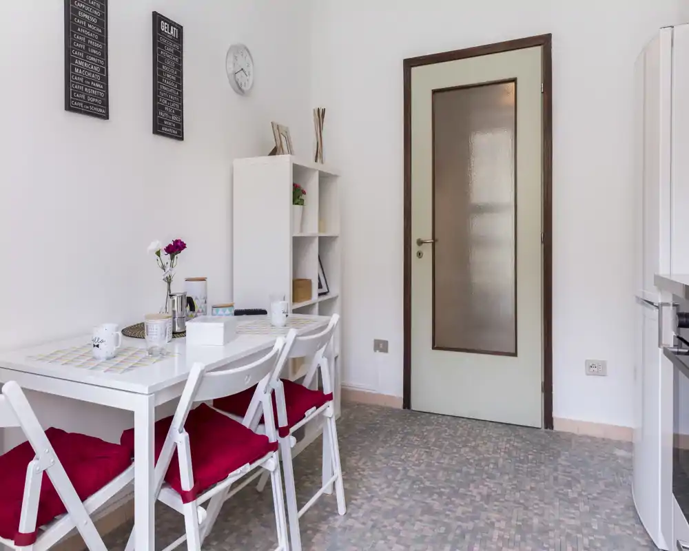 Bright, tidy dining nook with a white table and three chairs topped with red cushions, decorative placemats and small shelving — ideal for casual meals.