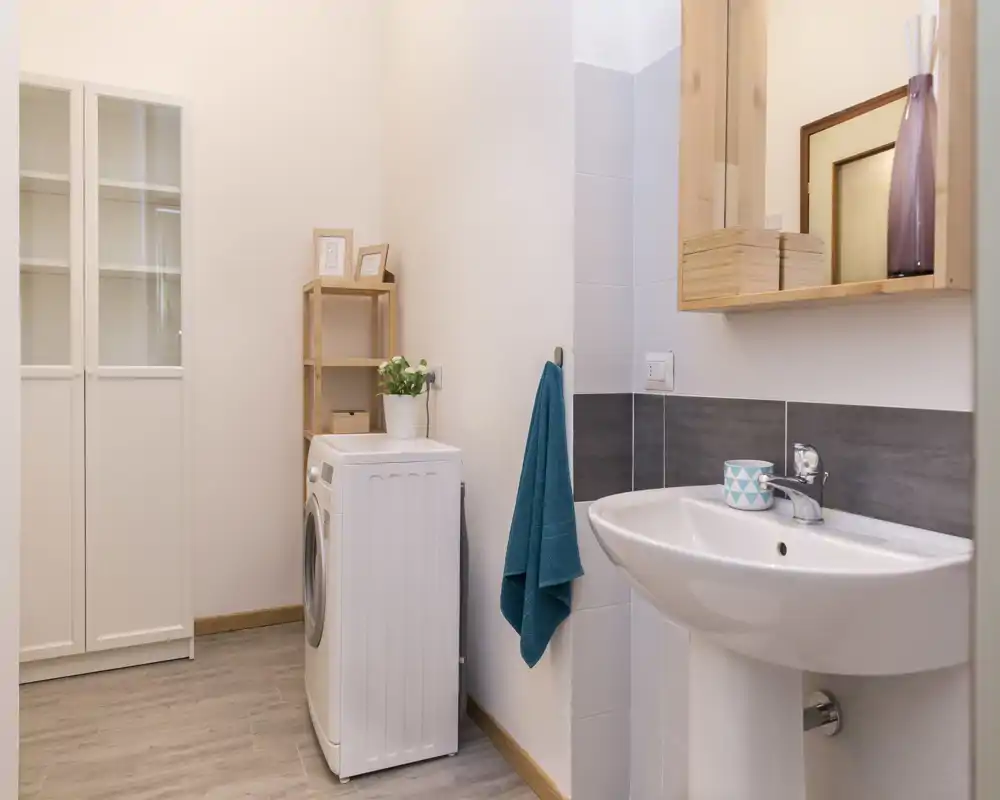 Modern, well-lit bathroom corner with a pedestal sink, mirror cabinet and a washing machine; neutral tiles and wooden accents create a clean, functional look.